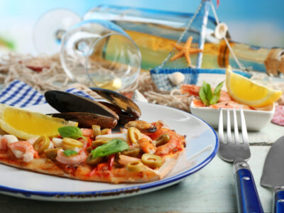 Pizza slice with seafood, red pepper, green olives and bottle of wine on wooden table