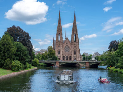 The Saint Paul Church of Strasbourg in France stands by the Ill river with twin spires reflected in calm water surrounded by green trees